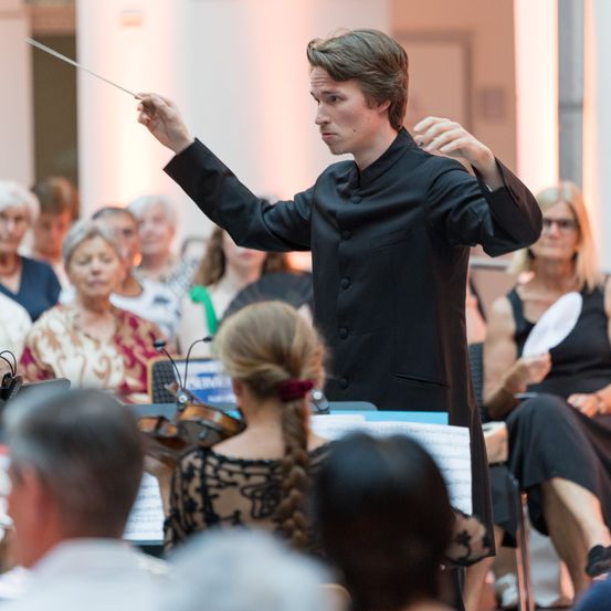 Ein junger Dirigent leitet ein Orchester, hält einen Taktstock vor einem sitzenden Publikum und einer Geigerin.