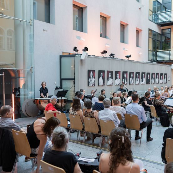 Ein Konzert mit einer Band und einem Orchester findet in einer Halle statt. Eine Frau steht mit einem Schlagzeug und einem Mikrofon auf der Bühne. Das Publikum sitzt in Stühlen vor der Bühne.
