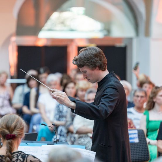 Ein Dirigent leitet eine Orchesteraufführung, steht vor einem Mikrofon und Notenblättern und wird von einem sitzenden Publikum beobachtet.