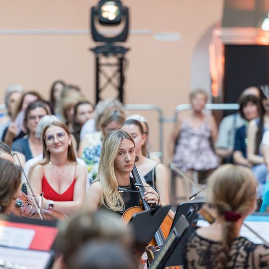 Eine Gruppe von Menschen beobachtet eine Frau, die in einem Konzert eine Geige spielt. Das Publikum ist verschwommen, und einige tragen Brillen. Die Bühne ist schwach beleuchtet.