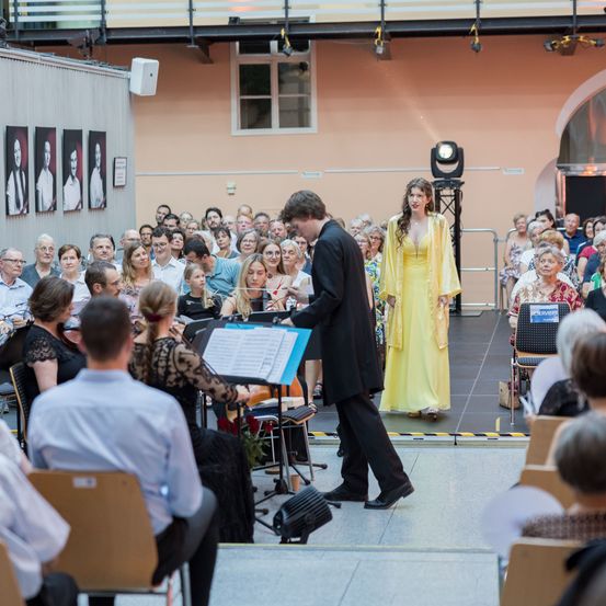 Ein Mann und eine Frau stehen auf der Bühne und führen vor einem Publikum auf. Die Frau im gelben Kleid steht neben dem Mann im schwarzen Anzug. Hinter ihnen befinden sich Musikinstrumente und ein Scheinwerfer.