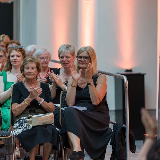 Eine Gruppe von Frauen sitzt in einem Raum, klatscht und lächelt. Sie tragen formelle Kleidung. Einige tragen Brillen und halten Taschen. Eine Frau in Schwarz steht und klatscht, während andere sitzen und zusehen.