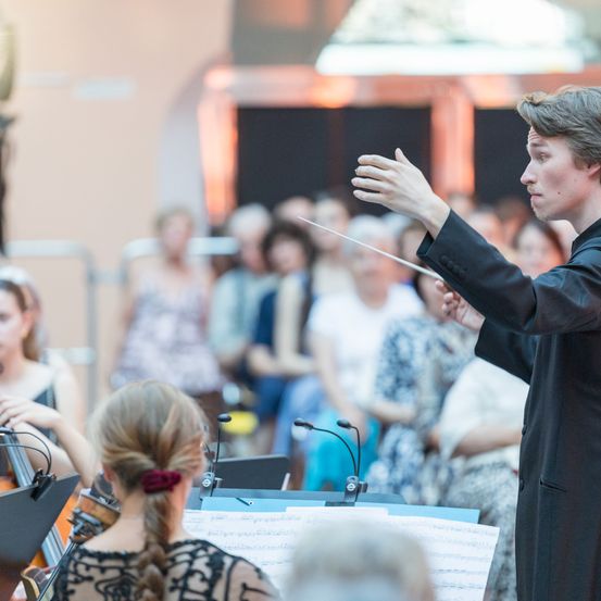 Ein junger Dirigent führt ein Orchester an, seine Hand erhoben, vor einem verschwommenen Publikum. Eine Cellistin sitzt in der Nähe, in ihr Instrument vertieft.