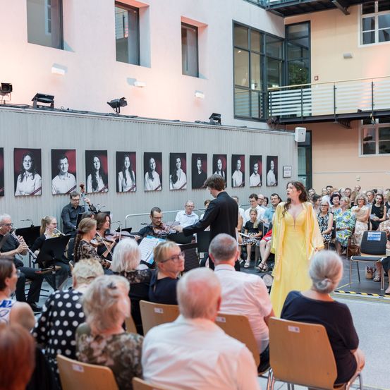 Ein Orchester spielt auf einer Bühne mit einer Frau in einem gelben Kleid vor dem Publikum. An der Wand hinter den Darstellern befinden sich Porträts von Personen.