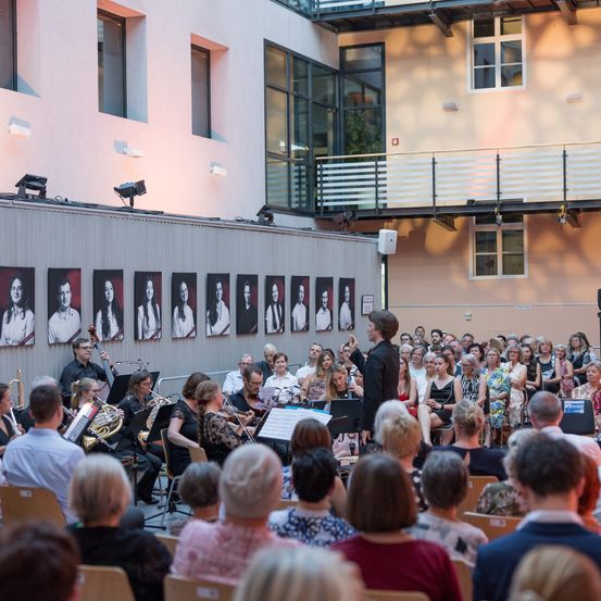 Ein Dirigent leitet ein Symphonieorchester, während eine vielfältige Menschenmenge im Publikum sitzt. Die Aufführung findet in einem modernen Gebäude mit vielen Fenstern und einem Balkon statt.