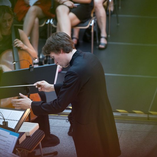 Ein Mann in einem schwarzen Anzug steht auf der Bühne, hält einen Taktstock und dirigiert eine Aufführung. Hinter ihm spielen Musiker, ein Cellist ist sichtbar. Das Publikum sitzt im Hintergrund.