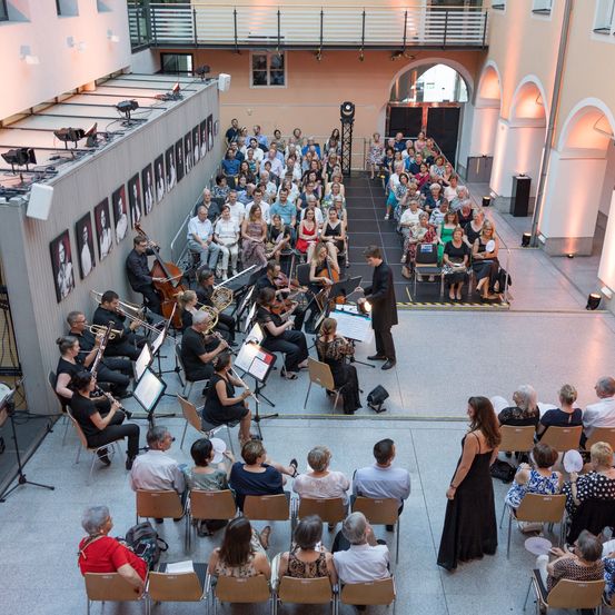 Ein klassisches Orchester spielt in einer modernen Location. Die Musiker spielen verschiedene Instrumente. Das Publikum sitzt in Stühlen und beobachtet die Aufführung.