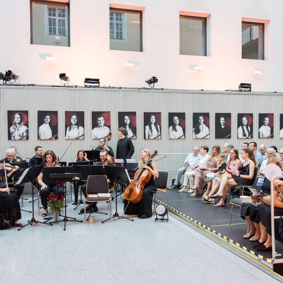 Ein Kammerorchester tritt auf der Bühne auf, mit Musikern, die verschiedene Instrumente spielen. Das Publikum sitzt vor der Bühne und beobachtet die Aufführung. Die Bühne ist mit gerahmten Porträts dekoriert.