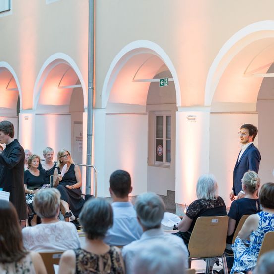 Eine Versammlung von Menschen in einem Raum mit gewölbten Decken. Ein Mann im Anzug spricht. Das Publikum sitzt, und eine Frau in Schwarz hält ein weißes Objekt.