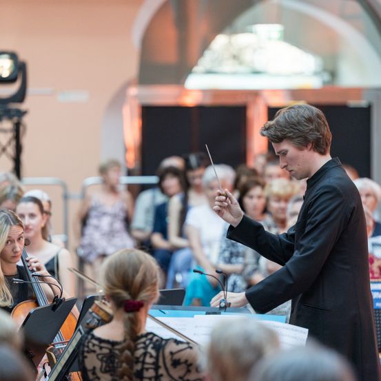 Ein junger Mann dirigiert ein Orchester in einem großen Saal mit einem Publikum im Hintergrund. Er hält einen Taktstock.
