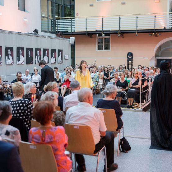 Eine Frau in einem gelben Kleid steht auf der Bühne vor einem Publikum. Sie wird von einem Mann in einem schwarzen Anzug begleitet. Das Publikum sitzt auf Stühlen, einige halten Kerzen. Der Raum hat große Fenster und Bilder an der Wand.