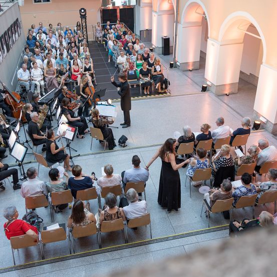 Ein Symphonieorchester tritt in einem großen Saal auf, das Publikum sitzt in Reihen. Der Dirigent leitet die Musiker, die verschiedene Instrumente spielen. Das Publikum hört aufmerksam zu.