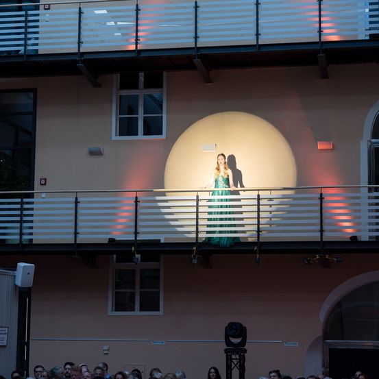 Eine Frau in einem grünen Kleid steht auf einem Balkon mit Geländer. Im Hintergrund befindet sich eine Wand mit Fenstern und roten Lichtern. Unten beobachtet eine Menschenmenge.