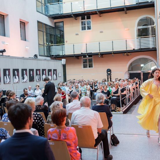 Ein Innenbereich mit Orchester und Publikum. Eine Frau in einem gelben Kleid geht auf die Bühne. Das Publikum sitzt in Reihen, einige stehen. Die Wände sind mit Porträts dekoriert.