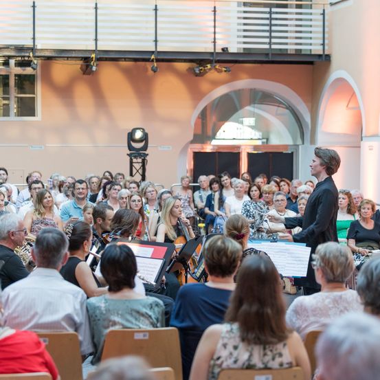 Ein Mann dirigiert ein Orchester in einer Halle mit einem großen Publikum. Das Publikum sitzt auf Stühlen vor den Musikern, die verschiedene Instrumente spielen. Die Halle hat eine hohe Decke mit dekorativen Bögen.