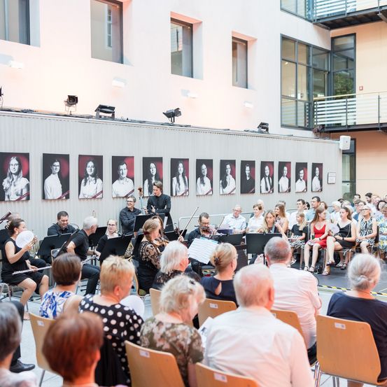 Ein klassisches Musikensemble spielt auf der Bühne mit Geigern, Cellisten und einem Dirigenten, während ein Publikum aus verschiedenen Personen davor sitzt. Hinter den Musikern befinden sich Porträtfotos verschiedener Musiker.