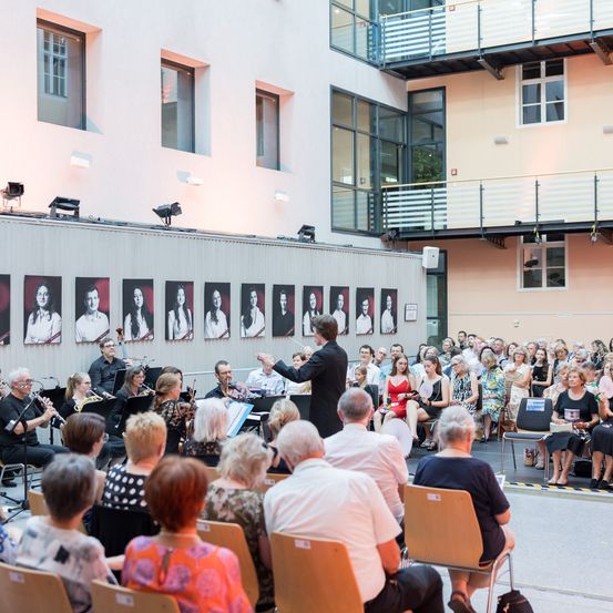 Ein Dirigent führt ein kleines Orchester an, während sie für ein großes Publikum in einem modernen Gebäude spielen. Das Publikum sitzt in Reihen, einige stehen. Die Bühne ist mit Porträts von Frauen geschmückt.