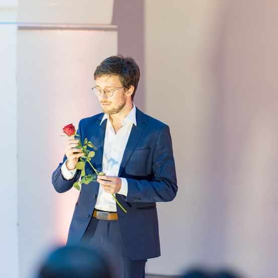 Ein Mann in einem blauen Anzug hält eine rote Rose. Er steht vor einer weißen Wand.