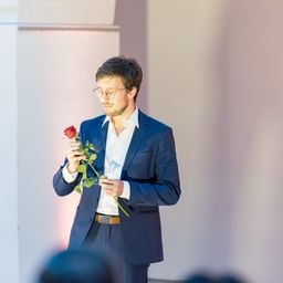 Ein Mann in einem blauen Anzug hält eine rote Rose. Er steht vor einer weißen Wand.