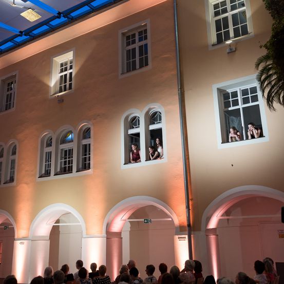 Eine Gruppe von Menschen schaut von den Fenstern eines Gebäudes auf eine Veranstaltung. Sie sitzen auf Bänken, einige von ihnen halten Tassen. Die Fenster haben bogenförmige Oberseiten.