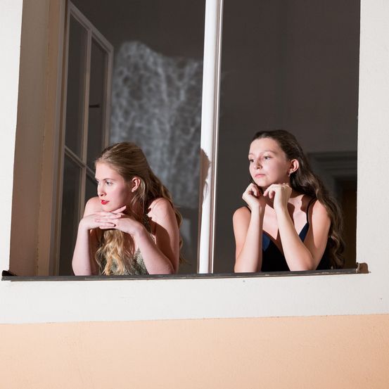 Zwei Frauen schauen aus einem Fenster. Eine hat die Hände auf der Brust und schaut nach rechts. Die andere hat die Hände am Kinn und schaut nach links.
