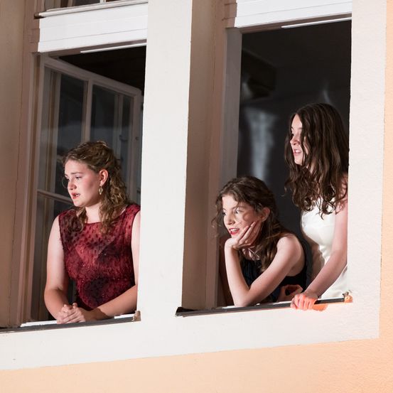 Drei Mädchen in Kleidern stehen in Fenstern. Ein Mädchen lächelt, ein anderes sieht nachdenklich aus und das dritte schaut nach draußen. Die Fenster haben weiße Rahmen.