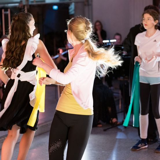 Drei Mädchen tanzen in einem Raum, eines mit einem gelb-schwarzen Kleid, eines mit einem rosa Top und schwarzen Leggings. Eine Person im Hintergrund spielt Flöte.