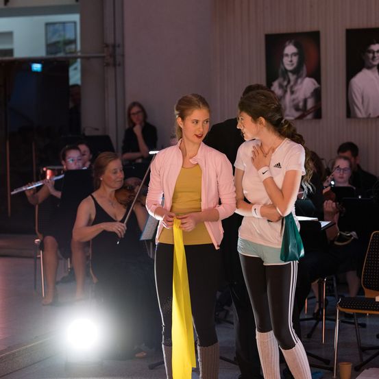 Zwei junge Frauen in Freizeitkleidung stehen vor einem Orchester, eine hält ein gelbes Band. Hinter ihnen spielen Musiker Instrumente, während andere beobachten. Der Raum ist schwach beleuchtet, mit einem fokussierten Licht auf den Darstellern.