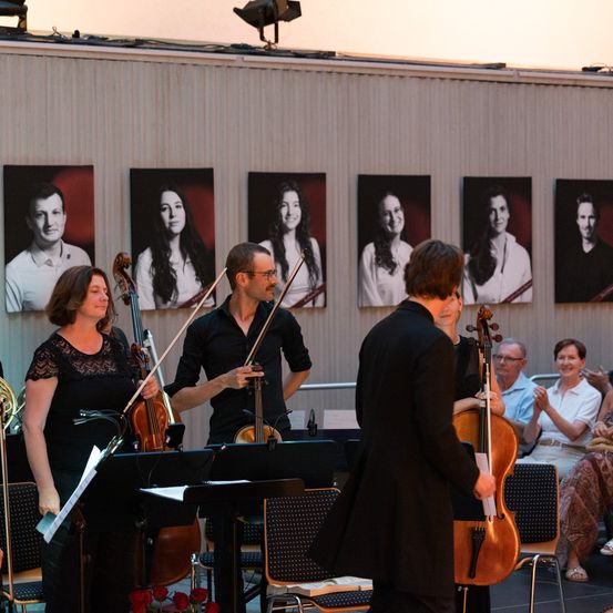 Eine Gruppe Musiker tritt auf der Bühne auf. Ein Mann steht mit Geige und Bogen, und eine Frau hält ein Cello. Hinter ihnen sind Fotos von Menschen an der Wand.