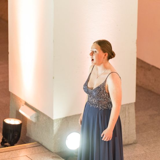 Eine Frau in einem blauen Kleid singt auf einer Bühne mit einer Säule und Lichtern hinter ihr.