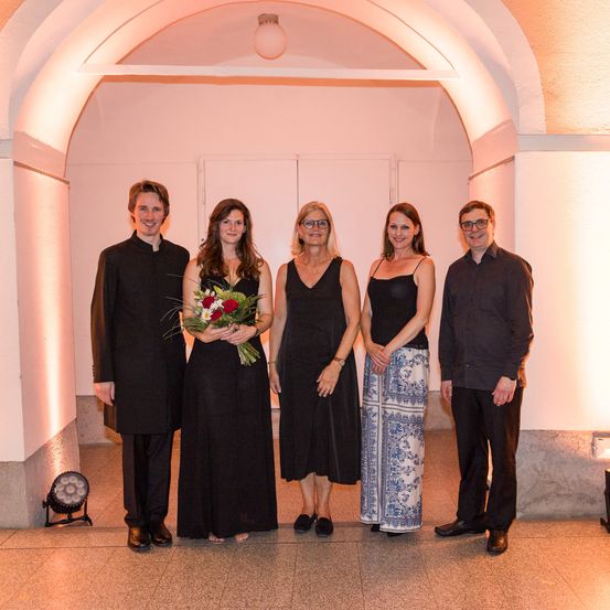 Fünf Personen, in Schwarz gekleidet, stehen unter einem Bogen mit rosa Licht. Die Frau in der Mitte hält Blumen. Sie scheinen ein Ereignis zu feiern.