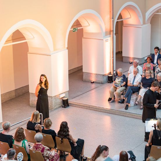 Eine Frau in einem schwarzen Kleid steht auf einer Bühne und spricht zu einem Publikum, das in Stühlen sitzt. Das Publikum besteht aus mehreren älteren Personen, von denen eine eine Geige spielt. Die Bühne hat eine gewölbte Struktur im Hintergrund.