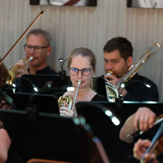Eine Band probt mit Trompeten und Posaunen, wobei eine Frau in der Mitte die Trompete spielt. Die Musiker tragen alle eine Brille.