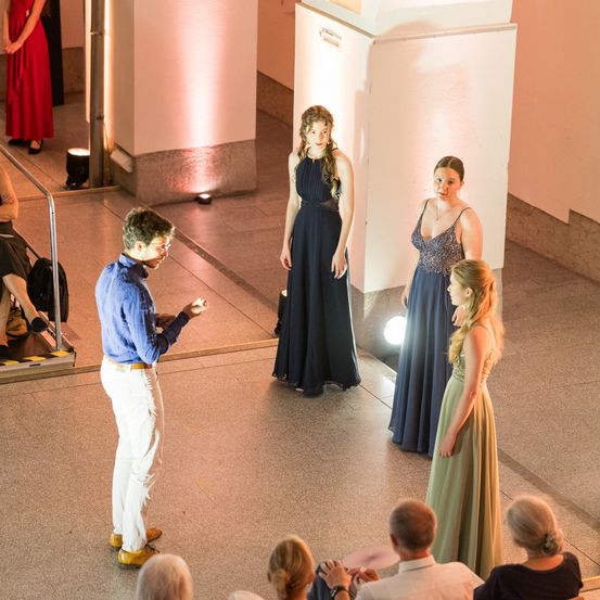 Ein Mann spricht ein Publikum an, während zwei Frauen in blauen Kleidern zuhören. Hinter ihm stehen zwei Frauen in Abendkleidern auf einer Bühne. Das Publikum beobachtet vom Balkon.