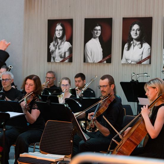 Eine Gruppe von Musikern, darunter Geiger und eine Cellistin, spielt in einem Innenraum. Sie sitzen auf Stühlen und spielen ihre Instrumente. Drei gerahmte Porträts hängen an der Wand hinter ihnen.