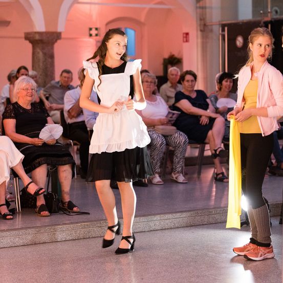 Zwei junge Frauen tanzen auf der Bühne, während mehrere ältere Frauen im Publikum zusehen.