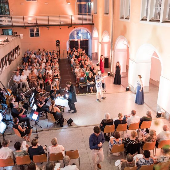 Ein Konzert findet in einem großen Saal statt, in dem das Publikum in Reihen sitzt. Die Musiker spielen auf der Bühne, und das Publikum beobachtet sie. Auf der Bühne stehen zwei Frauen, von denen eine ein Mikrofon hält.
