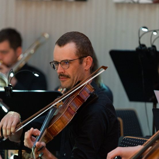 Ein Mann mit Brille spielt Geige in einem Musikensemble. Andere Musiker sind anwesend, darunter ein Mann, der Posaune spielt.