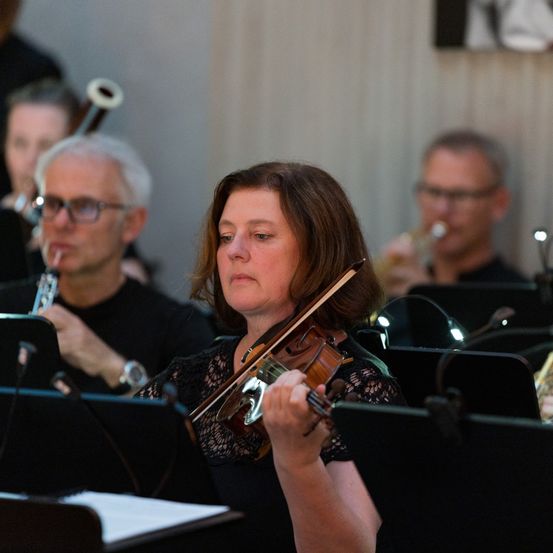 Eine Violinistin spielt in einem Orchester mit mehreren Musikern, einschließlich eines Trompeters. Das Orchester ist drinnen.