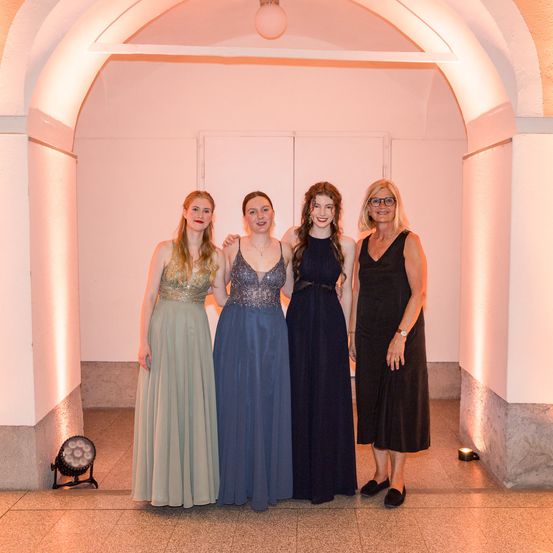 Vier Frauen in Abendkleidern posieren für ein Foto, zwei mit verbundenen Armen, in einem warm beleuchteten Raum.