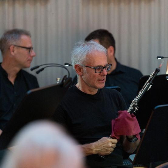 Ein älterer Mann in einem schwarzen T-Shirt spielt Klarinette, während zwei andere hinter ihm sitzen. Sie sind wahrscheinlich in einer Musikprobe oder Aufführung.