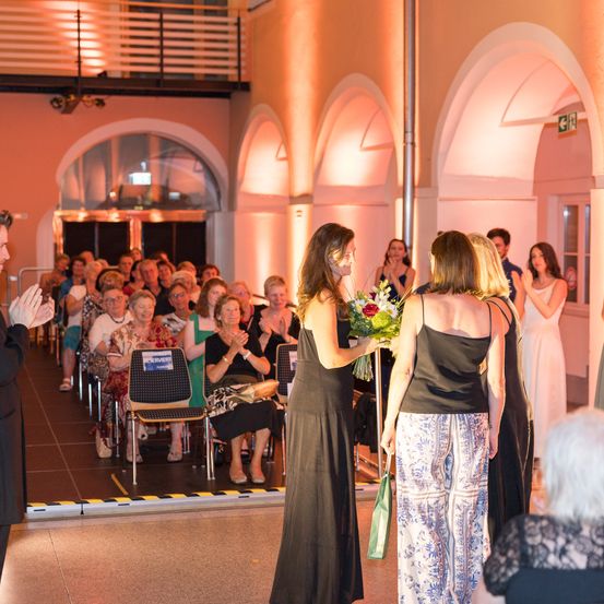 Eine Gruppe von Frauen steht in einer Innenumgebung, eine hält Blumen. Ein Mann klatscht vor ihnen. Das Publikum sitzt auf Stühlen und beobachtet. Der Raum hat gewölbte Fenster und eine rosa Beleuchtung.