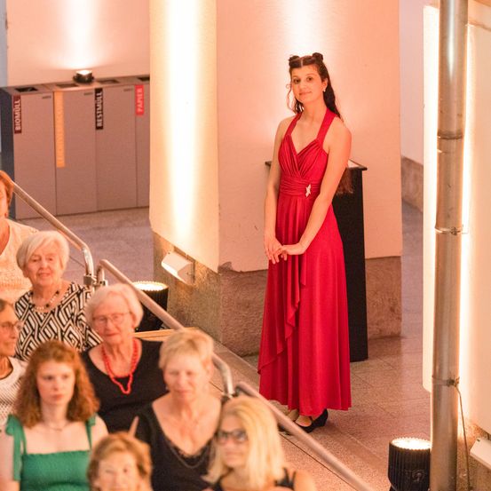 Eine Frau in einem roten Kleid steht in einem Theater und schaut auf ein Publikum älterer Frauen in verschiedenen farbigen Kleidern. Hinter ihr sind Mülleimer mit Etiketten zu sehen.