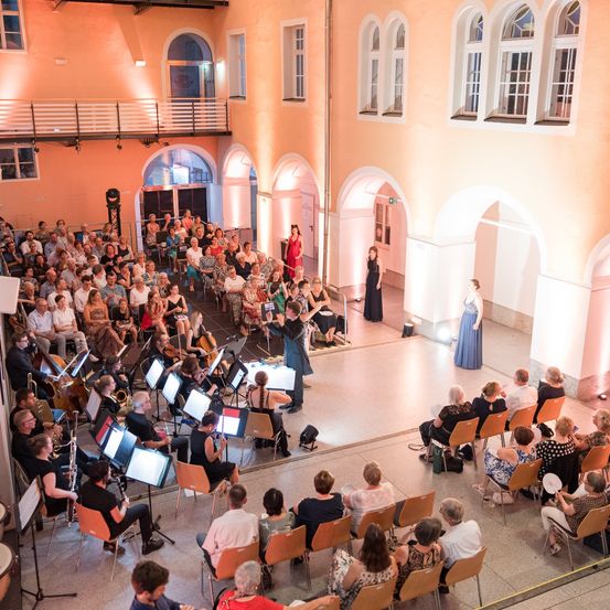 Ein Orchester spielt in einem Saal vor Publikum. Musiker sitzen auf der Bühne, während ein Dirigent davor steht. Einige Zuschauer stehen auf und klatschen.