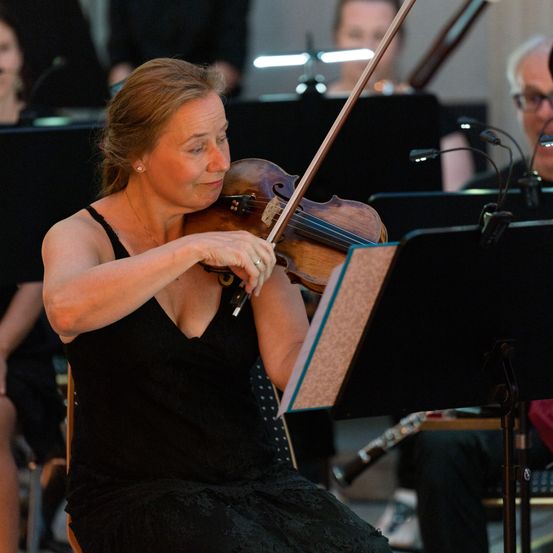 Eine Frau in einem schwarzen Kleid spielt Geige, umgeben von anderen Musikern. Der Fokus liegt auf ihrem intensiven Blick und der Geige in ihren Händen.