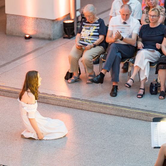 Eine Frau in einem weißen Kleid sitzt auf dem Boden und schaut in Richtung der sitzenden Personen. Einige tragen Brillen und Schuhe. Eine hält ein Buch. Rechts liegt ein Notenblatt.