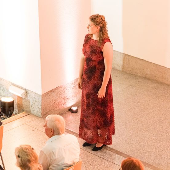 Eine Frau in einem roten Kleid steht vor einem sitzenden Publikum, Scheinwerfer beleuchten sie. Hinter ihr sind Wände rosa und Marmor, und der Boden ist gefliest.