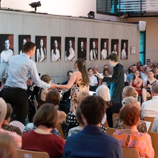 Eine Aufführung, bei der ein Mann und eine Frau auf der Bühne Hände schütteln. Das Publikum sieht von ihren Plätzen zu. Hinter ihnen hängen Bilder an der Wand.