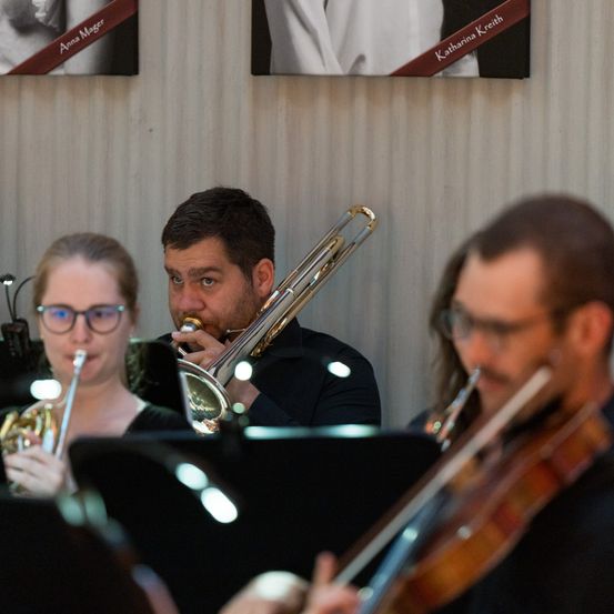 Ein Orchester spielt während eines Konzerts. Ein Mann spielt Posaune, eine Frau Trompete und ein Mann Geige. Hinter ihnen eine Wand mit gerahmten Porträts von Anna Mager und Katharina Kreith.