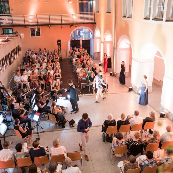 Ein klassisches Musikkonzert ist im Gange. Ein Orchester ist auf der Bühne, und zwei Frauen in Abendkleidern stehen in der Nähe. Das Publikum sitzt auf Stühlen vor der Bühne. Der Veranstaltungsort ist schwach beleuchtet.